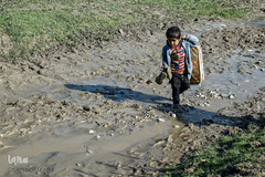 ابوالفضل با بدست گرفتن کفش‌های خود سعی بر رد شدن از گل و لای جاده را دارد؛ مسیرهای کوهستانی و خاکی روستا در زمان بارندگی و فصل سرما عبور و مرور را برای ساکنین این منطقه دشوار کرده است.
